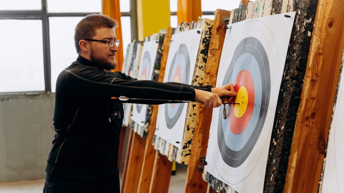 Man in glasses practicing archery indoors, pulling arrows from bullseye on target.